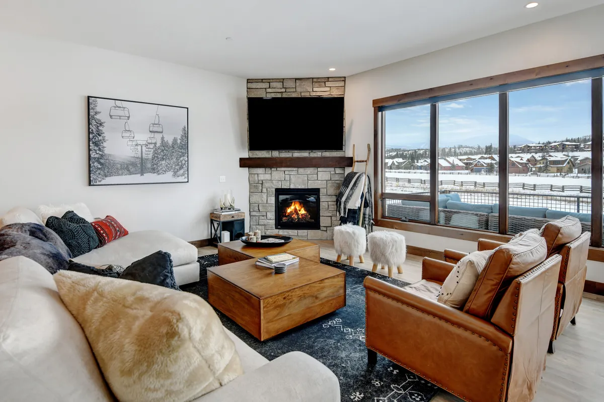 This cozy living area centers around a striking fireplace, with plush seating arranged for conversation or relaxation. Expansive windows let in abundant natural light and offer glimpses of snow-capped mountains, while a contemporary coffee table and tasteful artwork complete the welcoming scene.