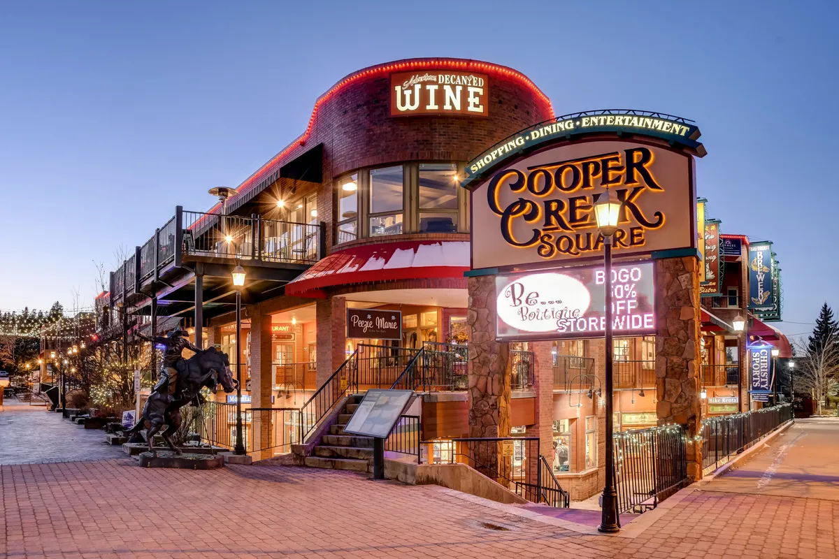 A lively exterior shot of Cooper Creek Square, featuring its iconic sign, a wine shop, inviting balcony stairs, and public art. The space is clearly designed for shopping, dining, entertainment, and social gatherings, capturing the downtown vibrancy and walkability.