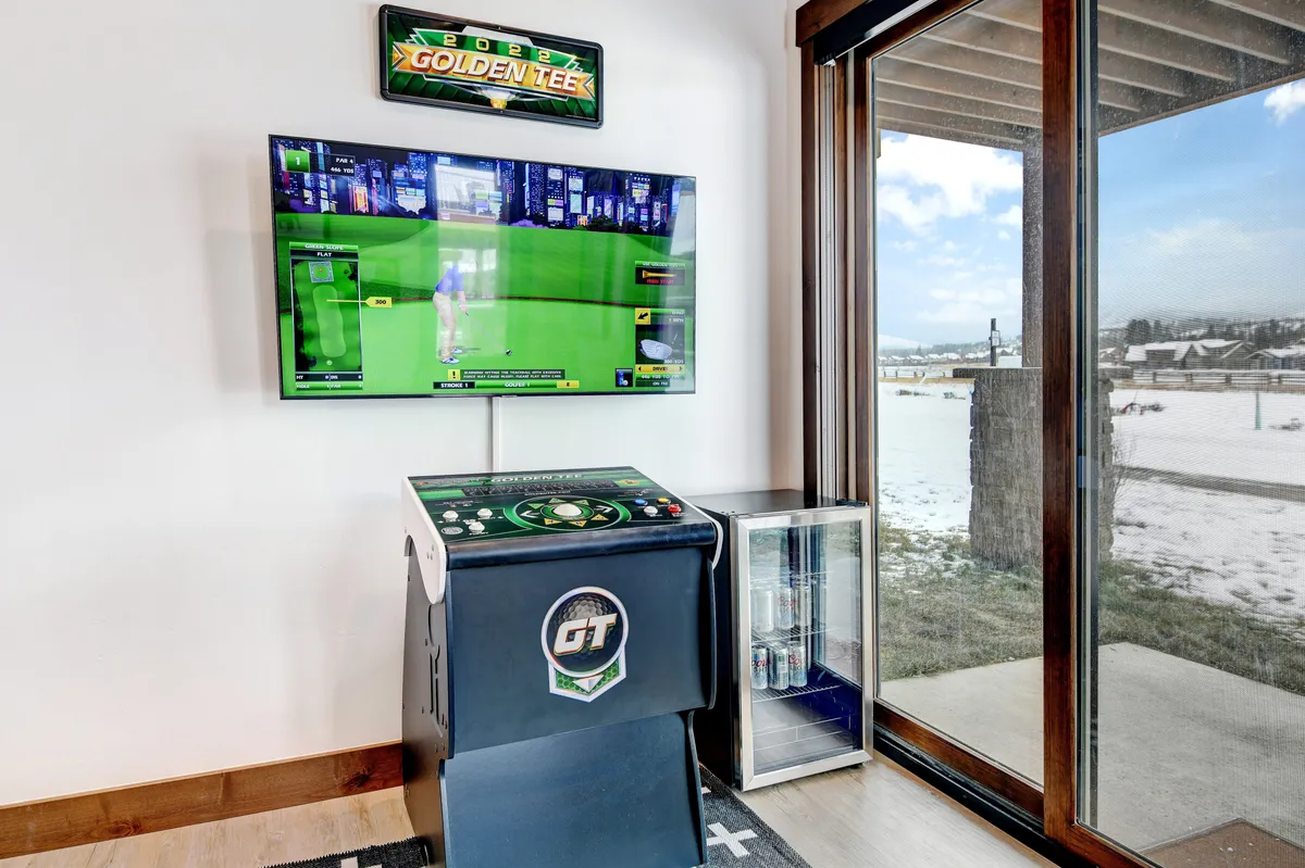A cozy corner of the game zone where the Golden Tee arcade game is set up beside a TV screen displaying a golf game. The area includes a glass door leading outside, providing both recreation and a glimpse of the outdoors.