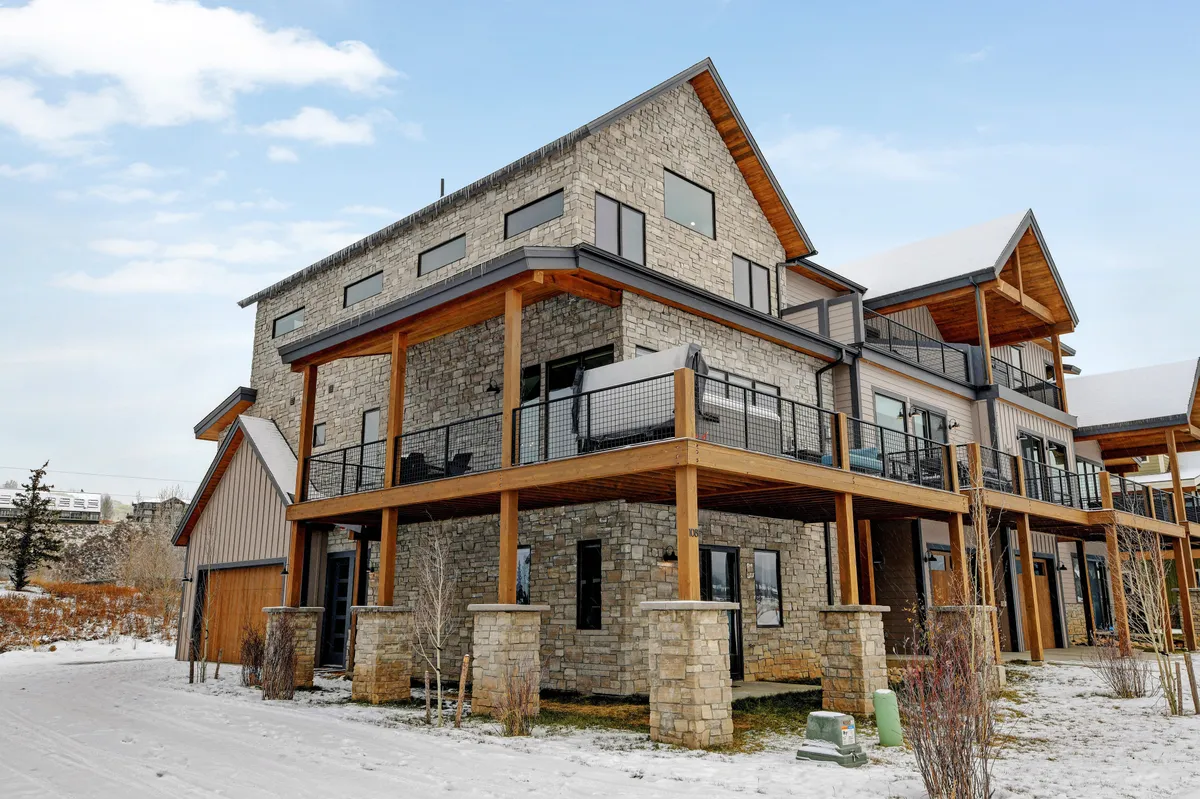 The image showcases the exterior of a luxurious mountain villa in Fraser, Colorado, featuring a stunning stone facade, elegant wooden beams, a spacious balcony, and expansive windows. The scene is set at sunset, casting a warm glow over the home and highlighting the inviting balcony, perfect for outdoor gatherings or soaking in breathtaking mountain views. Snow-dusted surroundings hint at nearby winter adventures and activities.