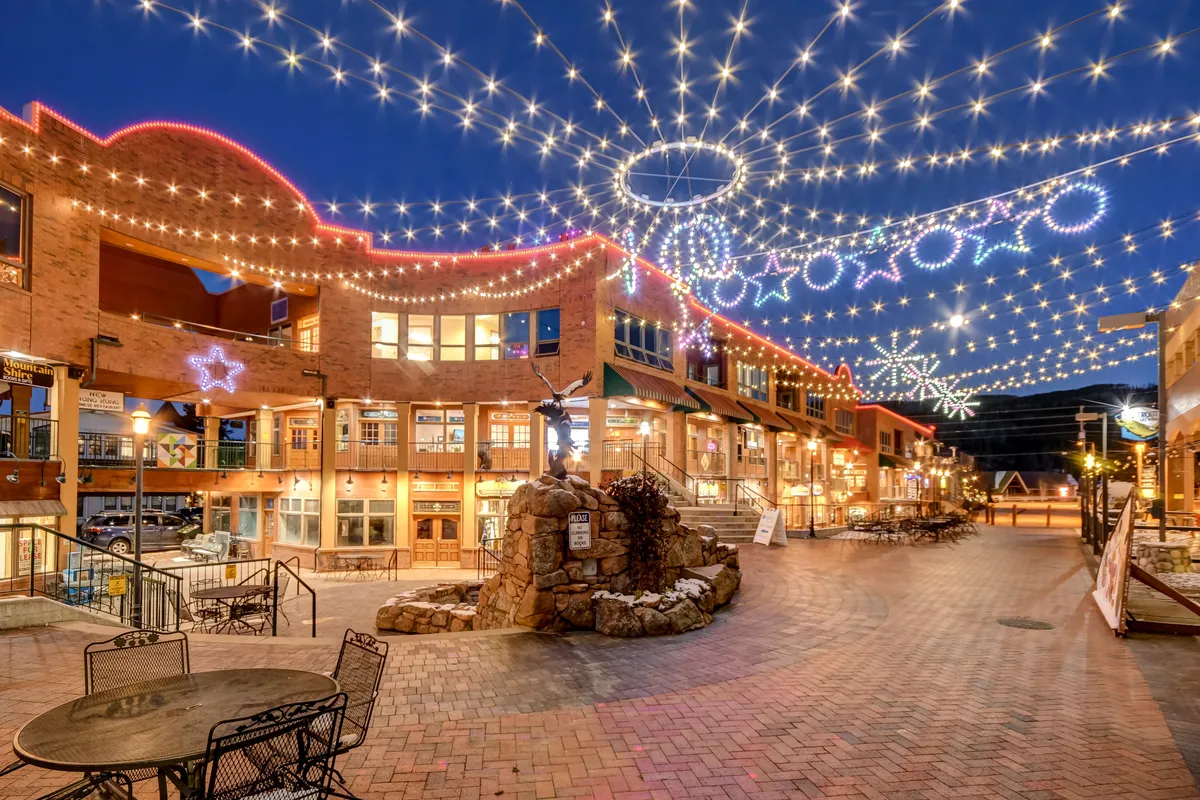 A charming outdoor plaza scene illuminated by decorative string lights and star decorations. The area features a rock fountain, ample outdoor seating, and a welcoming atmosphere, suggesting a lively community hub or a place for evening relaxation.
