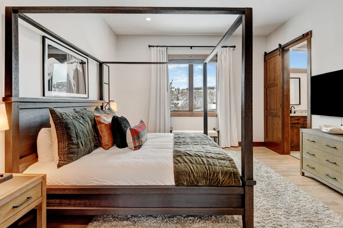 A serene bedroom featuring a four-poster bed with plush bedding, a large window offering a scenic view, decorative pillows, and matching nightstands. The soft natural light creates a tranquil ambiance perfect for rest.