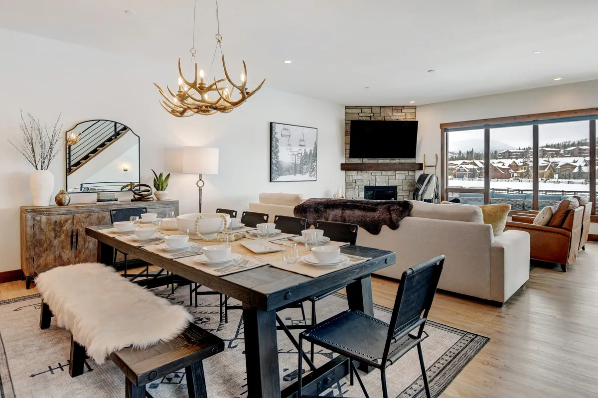 This image features a spacious open-concept dining and living area. The dining table is elegantly set beneath a chandelier, while a sofa area offers additional comfortable seating. A large fireplace serves as a stunning focal point, and sliding glass doors frame breathtaking mountain views, bathing the space in natural light.