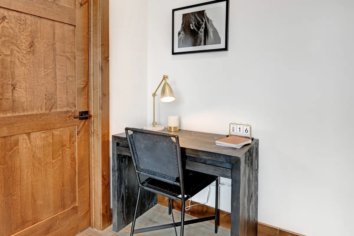 A serene workspace featuring a wooden desk equipped with a table lamp, framed artwork, and carefully arranged desk accessories. The desk is positioned near a wooden door, providing a quiet atmosphere ideal for focused work or creative pursuits.