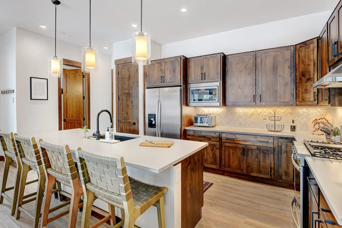 A modern kitchen is showcased here, featuring a granite-topped island surrounded by bar stools, wooden cabinetry, and stylish lighting fixtures. Premium appliances are visible, creating an inviting space for cooking, entertaining, and sharing casual meals or après-ski snacks.