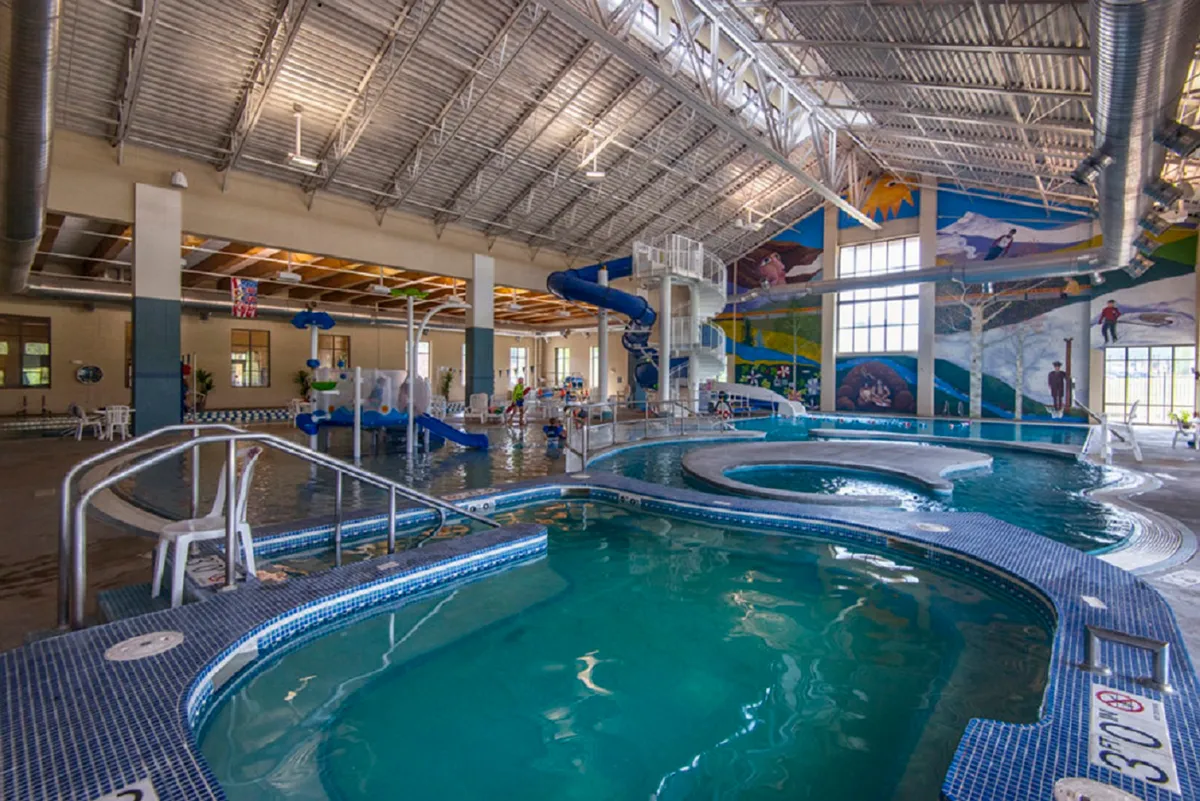 An interior view of the Grand Park Rec Center's aquatic area, complete with waterslides, swimming pools, and a vibrant wall mural. The scene is bright and family-friendly, suggesting a space for fun and recreation for all ages.