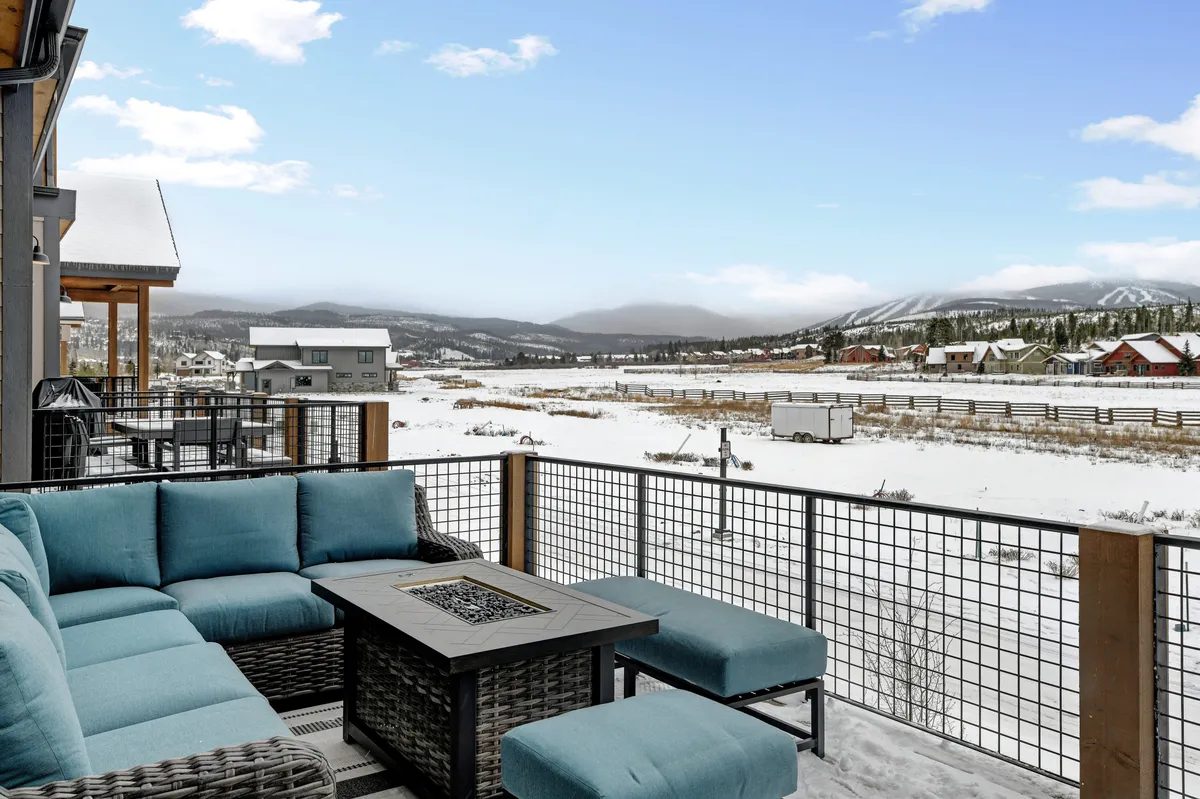An inviting patio lounge features modern blue outdoor seating and a fire pit table, all framed by a snow-covered landscape and distant mountain peaks. The setting is perfect for socializing, enjoying mountain air, and stargazing as evening falls.