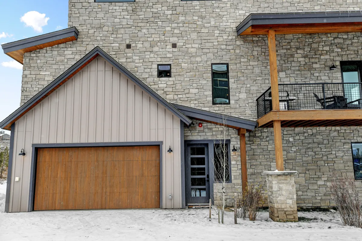This exterior image showcases the stone facade of Villa 108, featuring a welcoming wooden garage door, large windows, and a balcony area. The snow-dusted yard hints at winter fun, while the cozy architecture invites families to gather both inside and out.