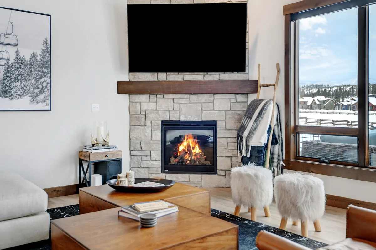 A welcoming living area with a cozy fireplace, large windows revealing mountain views, a coffee table, and artwork. The inviting ambiance is enhanced by soft seating and the glow of the fire, perfect for unwinding after a productive work session or enjoying a warm beverage while admiring the scenery.